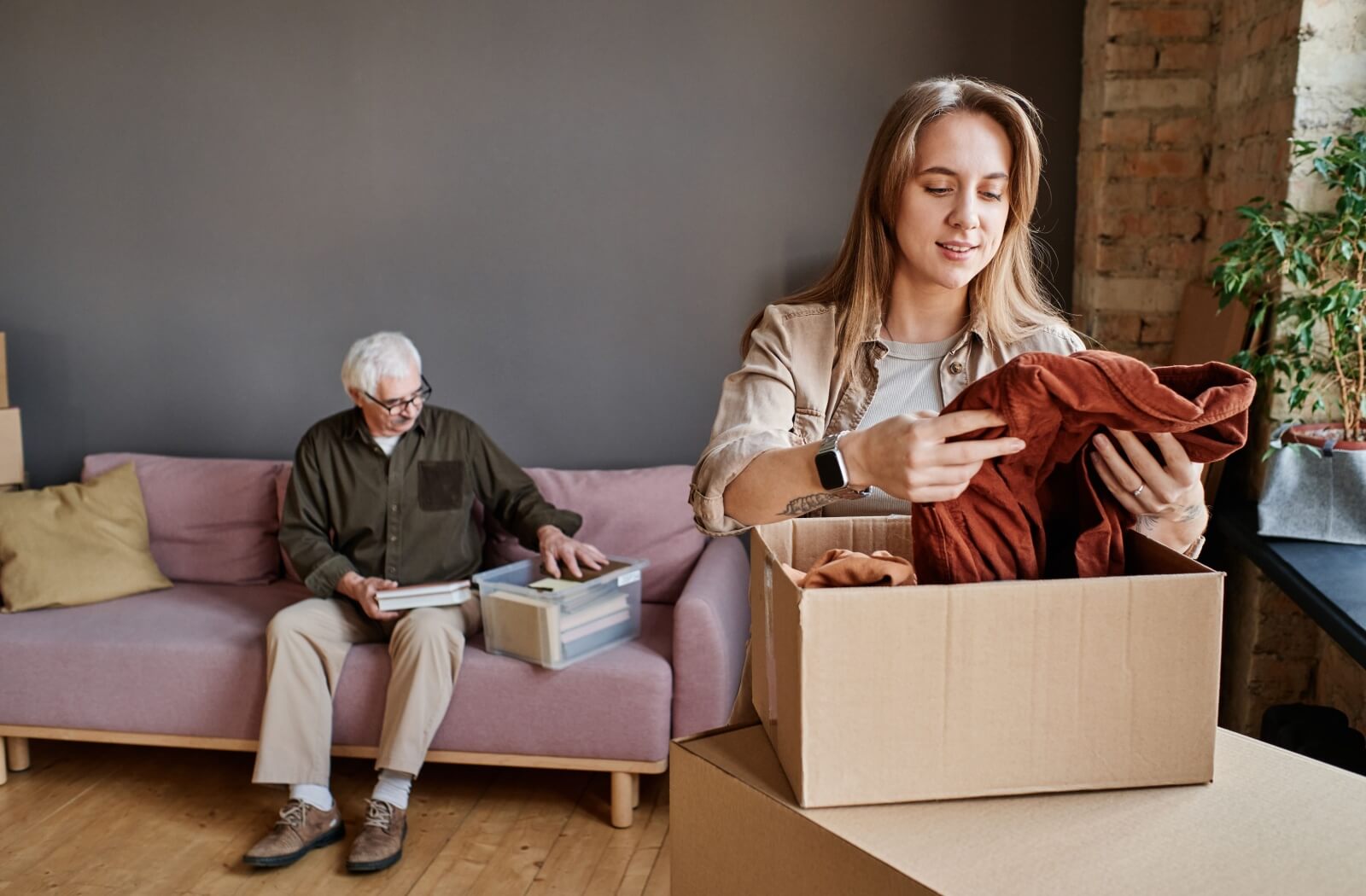 Person packing clothes in a box while older adult organizes books nearby during home move transition