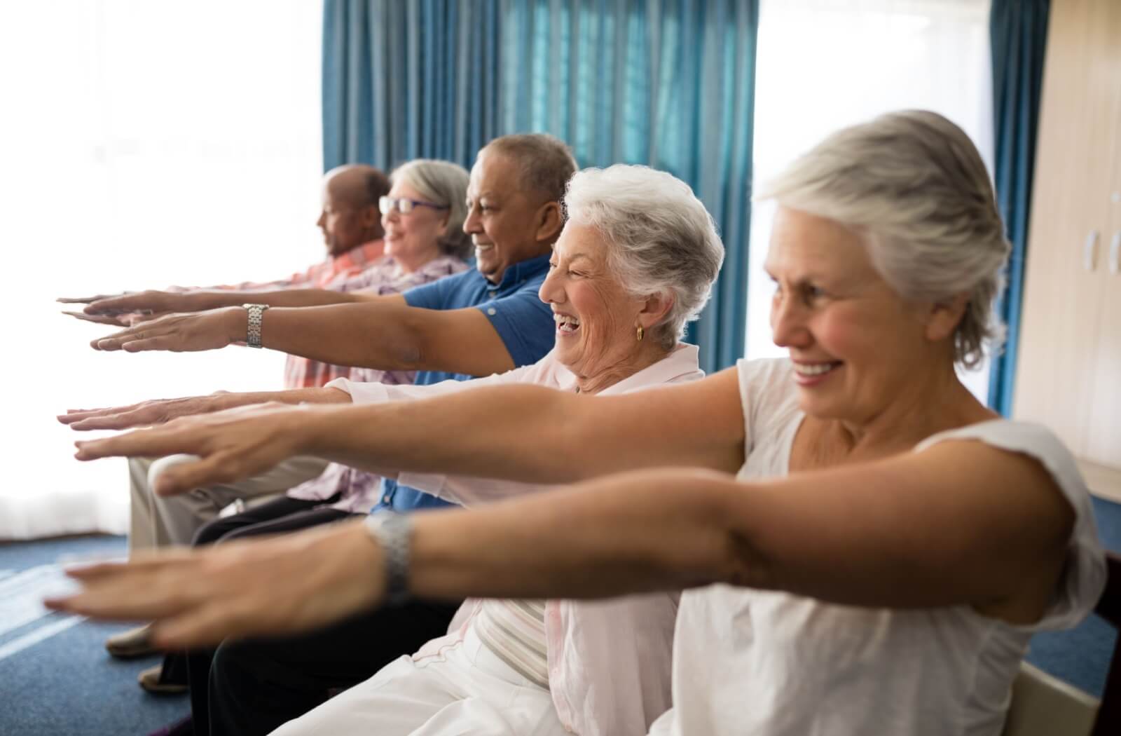 Group of smiling seniors seated indoors in a row, arms extended forward, doing exercises in natural sunlight