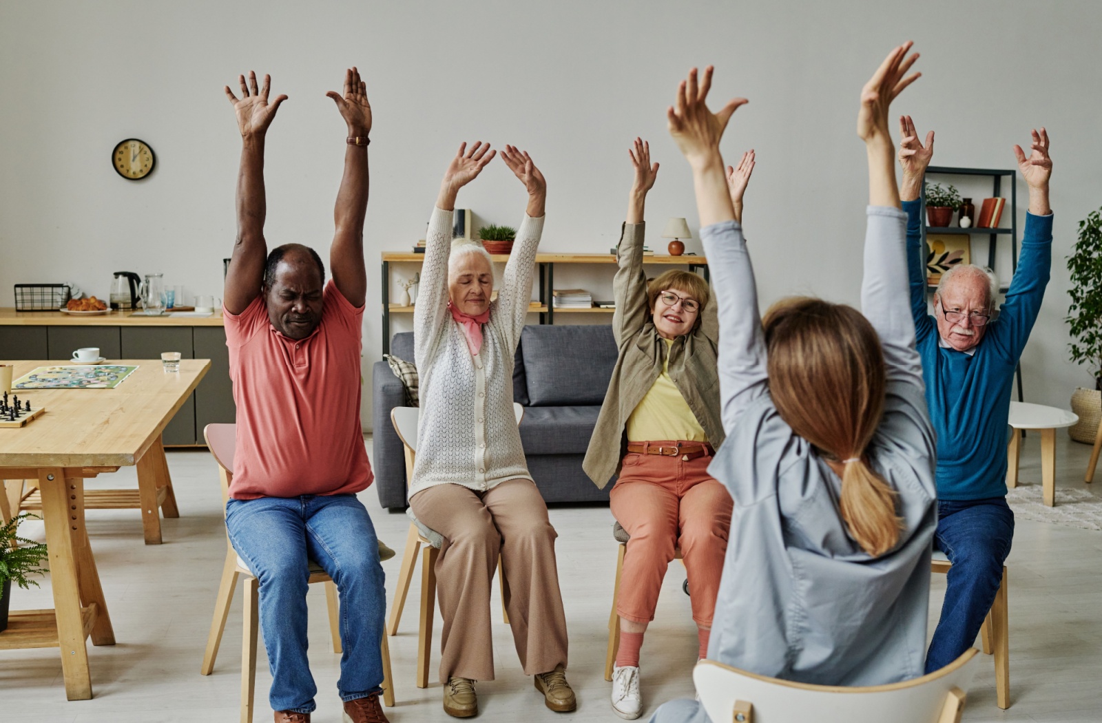 Seniors seated in a bright living space, stretching arms overhead with an instructor guiding them