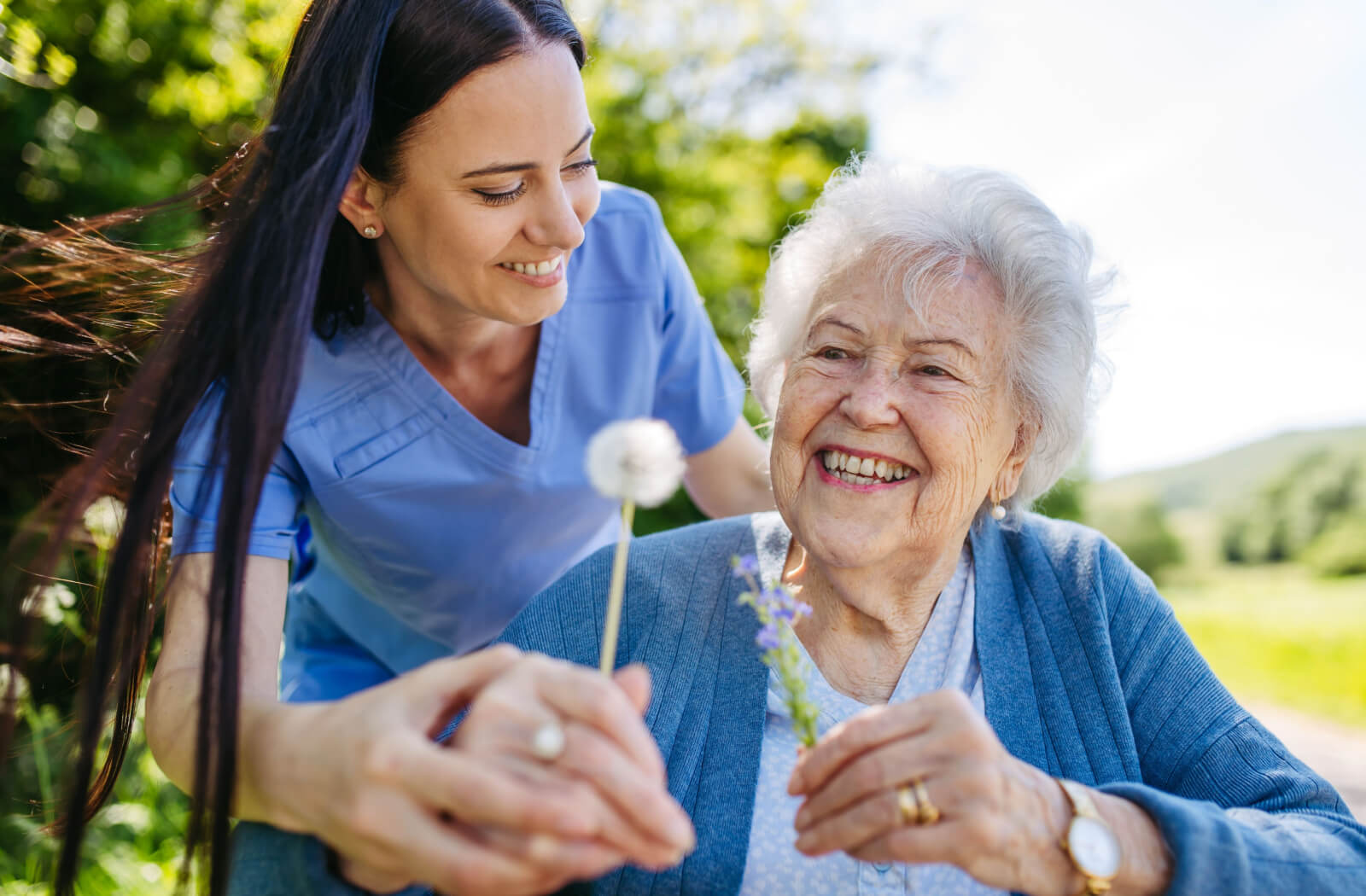 A caregiver handing a smiling older adult a dandelion while on a walk outside memory care.