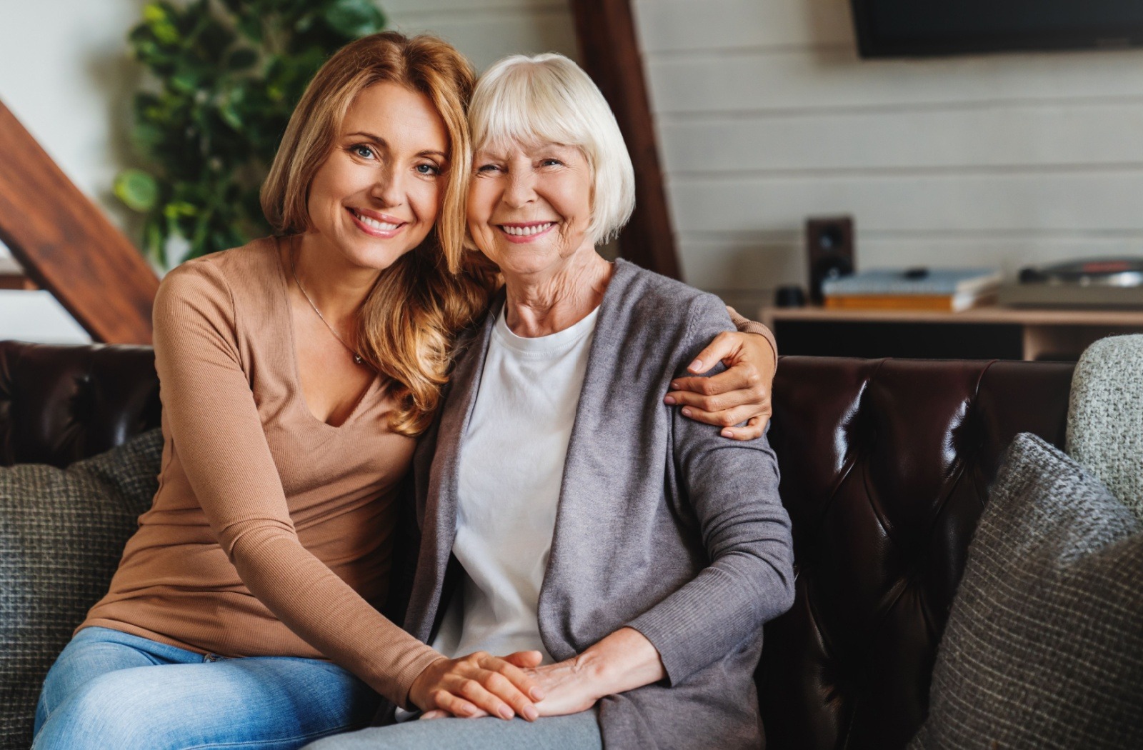A smiling adult child puts their arm around their older parent while sitting together on a leather couch